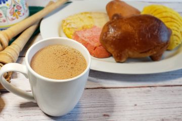 A cup of Authentic Mexican Hot Chocolate in 10 Minutes sits in the forefront. A plate of assorted Mexican cookies sit in the background, waiting to be dunked in this warm beverage.