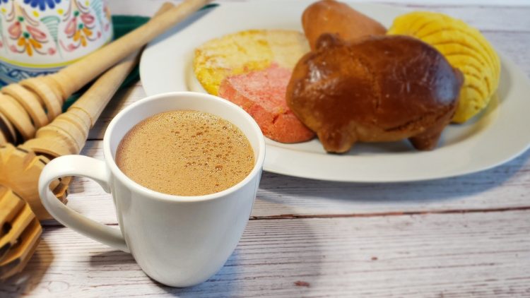 A cup of Authentic Mexican Hot Chocolate in 10 Minutes sits in the forefront. A plate of assorted Mexican cookies sit in the background, waiting to be dunked in this warm beverage.