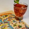 A refreshing glass of the Ultimate Smitten Mitten Cocktail sits atop a retro map of Michigan postcard. The drink is topped with a sprig of fresh mint.