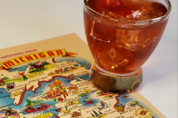 A refreshing glass of the Ultimate Smitten Mitten Cocktail sits atop a retro map of Michigan postcard. The drink is topped with a sprig of fresh mint.