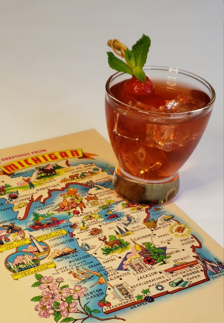 A refreshing glass of the Ultimate Smitten Mitten Cocktail sits atop a retro map of Michigan postcard. The drink is topped with a sprig of fresh mint.