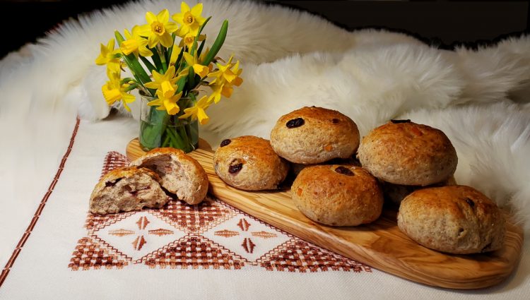 Stacked on top of a rustic wooden cutting board, sits a neat stack of Easy Nordic Rye Rolls in 3 Hours. The board rests upon a faux polar bear fur and nordic print linens. Vibrant spring flowers give the appearance of warmer weather breaking.