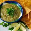 A bowl of Bright and Smoky Salsa Verde is surrounded by fresh lime wedges, cilantro and crisp corn tortilla chips.