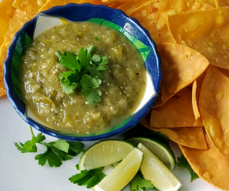 A bowl of Bright and Smoky Salsa Verde is surrounded by fresh lime wedges, cilantro and crisp corn tortilla chips.