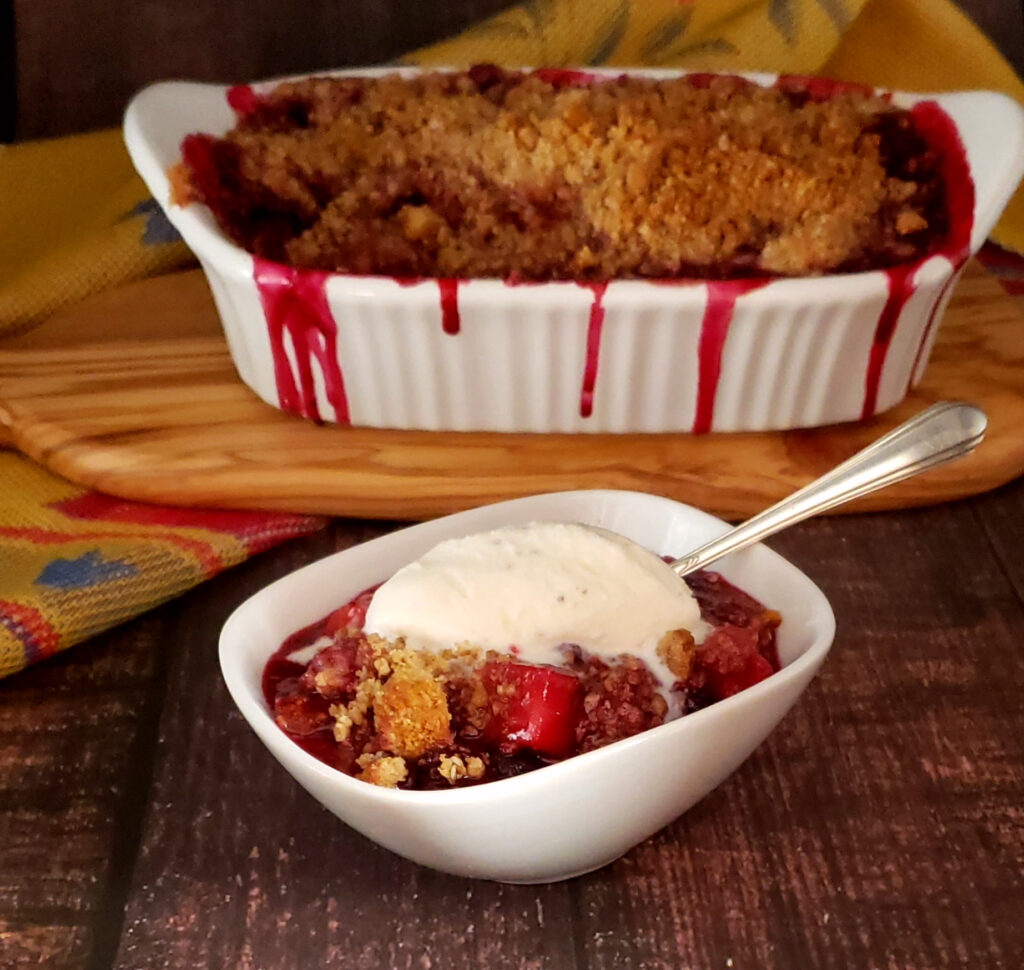 A bubbly hot baking dish full of Easy Apple Bumbleberry Cherry Crisp in 90 Minutes is packed with fruit and overflowing. A scoop of vanilla ice cream melts into the fruit and crisp topping. 