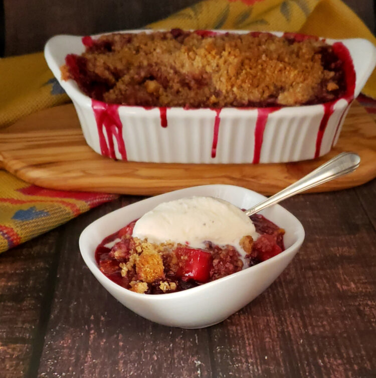 A bubbly hot baking dish full of Easy Apple Bumbleberry Cherry Crisp in 90 Minutes is packed with fruit and overflowing. A scoop of vanilla ice cream melts into the fruit and crisp topping.