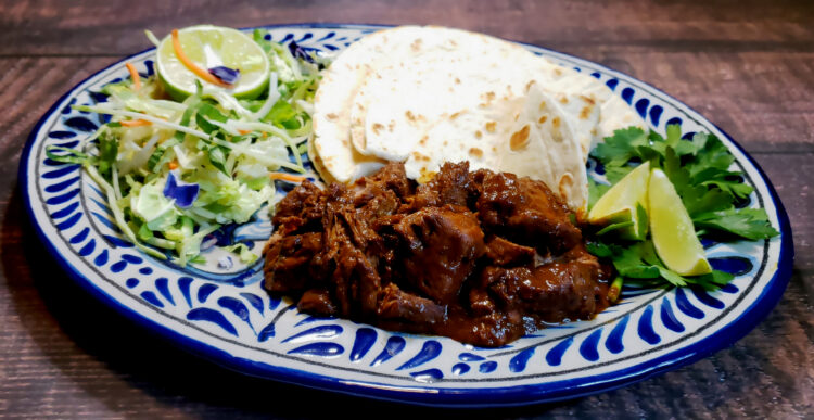 A beautiful Mexican-style plate of Delicious Pork and Prunes in Mole is served with a side of fresh greens, tortillas, a wedge of lime and cilantro.