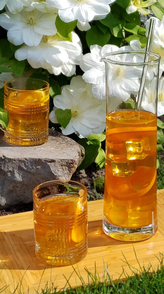 A tall pitcher of 3-Step Bless Your Heart Sweet Tea and two glasses at the ready catch the sunlight on a warm summer day. A clematis at the peak of its blooming season provides the backdrop for this beautiful, golden like sunshine beverage.