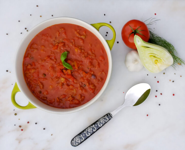 A beautiful pot of Maria's Best Spicy Sicilian Pasta Sauce sits on a marble board. Fresh fennel, tomatoes, garlic, onions and pepper corns are pictured next to the finished pot of sauce.