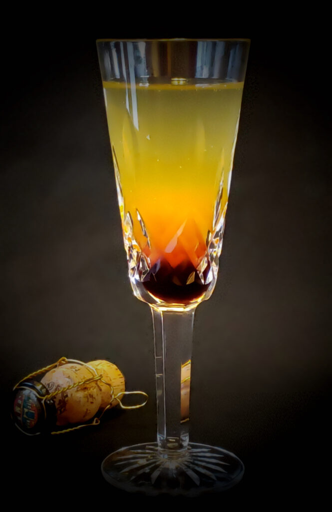 This image for Simple 3 Ingredient Tart Cherry Peach Bellini features a champagne flute with three distinct layers. The bottom of the glass has a layer of dark red cherry simple syrup. A bright layer of sunset orange peach nectar floats on top of the cherry layer. The cocktail is finished with a light layer of prosecco on top.