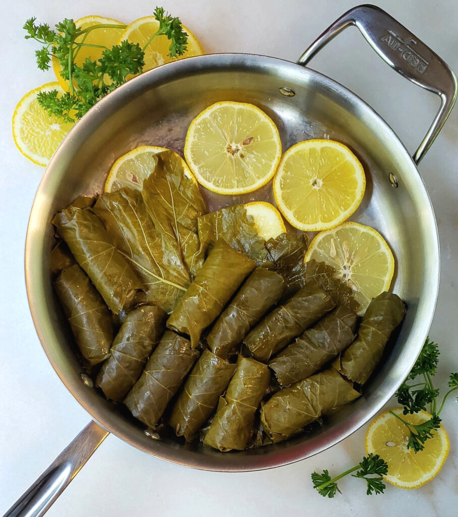 A pan is lined with lemons and grape leaves. Savory Tart Cherry Dolmas are lined up neatly in the pan - ready to be steamed.