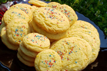 A large stand plate of Easy Mascarpone Orange Drop Cookies in 10 Minutes are nestled among flowers in a garden.