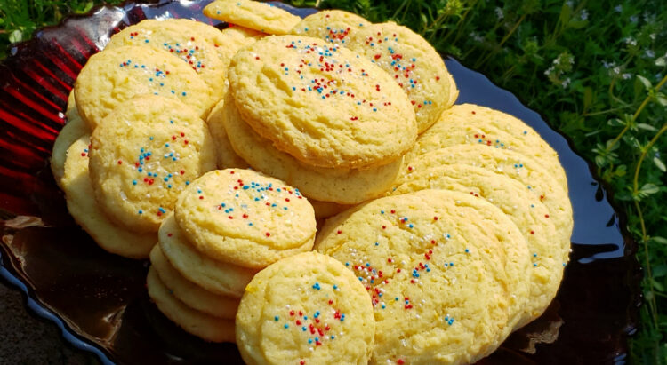 A large stand plate of Easy Mascarpone Orange Drop Cookies in 10 Minutes are nestled among flowers in a garden.