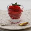 Easy Tart Cherry & Prosecco Sorbet. A simple and small glass of bright red tart cherry sorbet finished with a single sprig of mint sits upon a plate with a delicate pattern like lace. The white plate rests upon an antique, white linen.