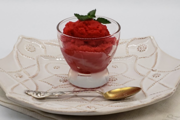 Easy Tart Cherry & Prosecco Sorbet. A simple and small glass of bright red tart cherry sorbet finished with a single sprig of mint sits upon a plate with a delicate pattern like lace. The white plate rests upon an antique, white linen.