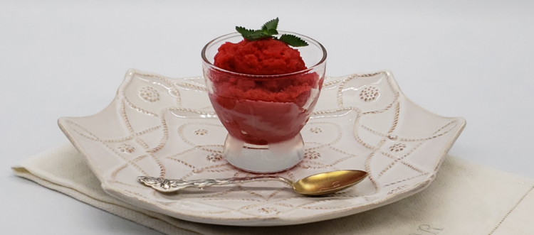 Easy Tart Cherry & Prosecco Sorbet. A simple and small glass of bright red tart cherry sorbet finished with a single sprig of mint sits upon a plate with a delicate pattern like lace. The white plate rests upon an antique, white linen.