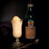 A dark, moody setting. bottle of Fernet Branca sits off to the right in the shadow. A Victorian style cordial glass holds a generous portion of Bold 6-Ingredient Fernet Branca Ice Cream. The color of the ice cream denotes hints of coffee and honey. A delicate, antique spoon rests across the plated dessert.