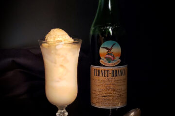 A dark, moody setting. bottle of Fernet Branca sits off to the right in the shadow. A Victorian style cordial glass holds a generous portion of Bold 6-Ingredient Fernet Branca Ice Cream. The color of the ice cream denotes hints of coffee and honey. A delicate, antique spoon rests across the plated dessert.