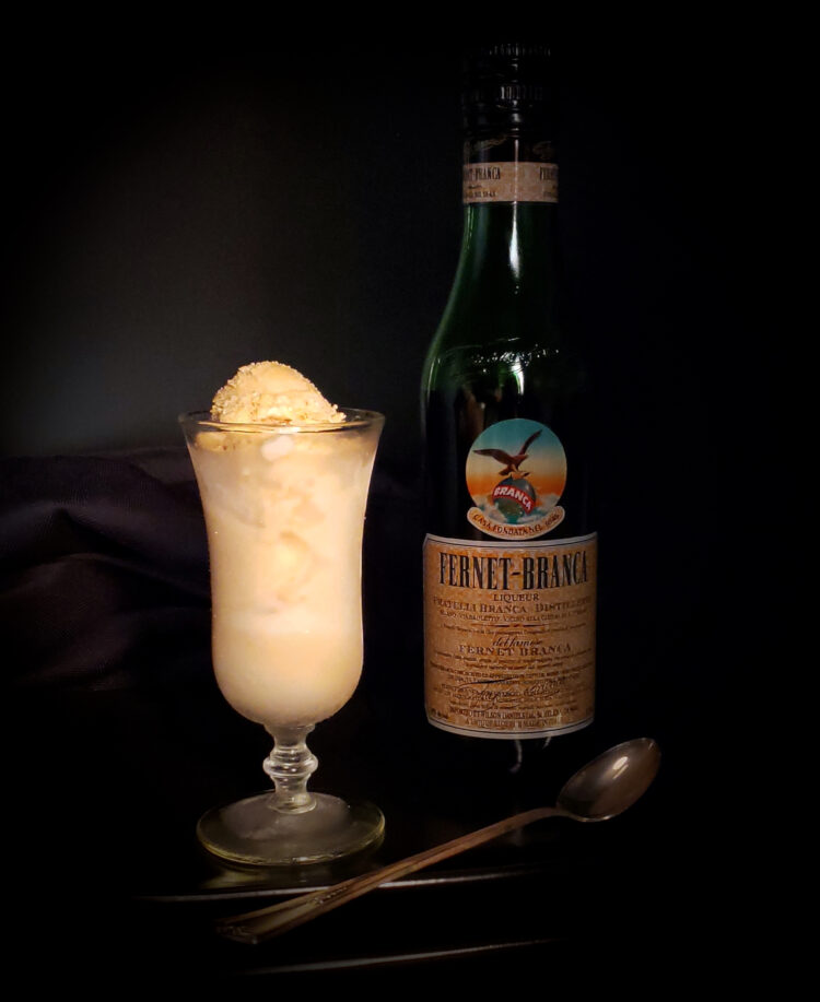 A dark, moody setting. bottle of Fernet Branca sits off to the right in the shadow. A Victorian style cordial glass holds a generous portion of Bold 6-Ingredient Fernet Branca Ice Cream. The color of the ice cream denotes hints of coffee and honey. A delicate, antique spoon rests across the plated dessert.