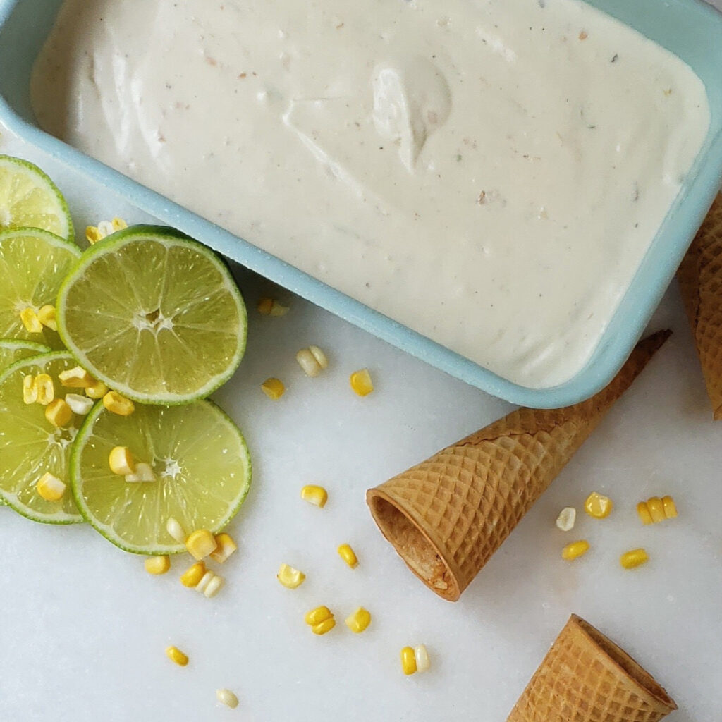 A fresh churned container of Playful 6-Ingredient Mexican Street Corn Gelato is featured on a cool marble slab. Fresh limes, corn kernels and ice cream cones are playfully placed around the container of ice cream.