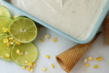 A fresh churned container of Playful 6-Ingredient Mexican Street Corn Gelato is featured on a cool marble slab. Fresh limes, corn kernels and ice cream cones are playfully placed around the container of ice cream.
