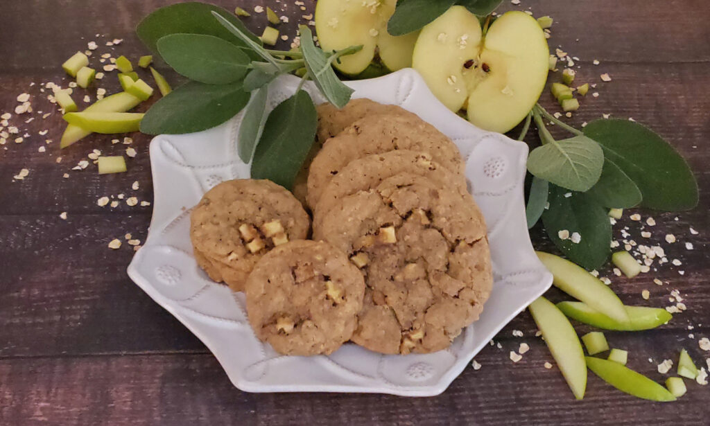 A rustic wood board holds a small, textured plate of Irresistible Apple Sage Oatmeal Cookies. Fresh sprigs of sage and oats frame the plate of cookies. A granny smith apple is sliced in half to expose the core and seeds. The image evokes the feel of crisp fall days, and warm apple pie.
