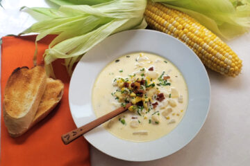 A piping hot bowl of Easy 30-minute corn chowder sits in the center of the frame. Crispy pancetta and fresh cut chives garnish the bowl. A rustic wooden spoon stands ready to dig in. Toasted fresh bread rests on a napkin to the left of the soup. A partially shucked ear of corn reveals the sweet summer vegetable and frames the picture.