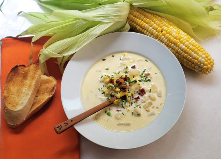A piping hot bowl of Easy 30-minute corn chowder sits in the center of the frame. Crispy pancetta and fresh cut chives garnish the bowl. A rustic wooden spoon stands ready to dig in. Toasted fresh bread rests on a napkin to the left of the soup. A partially shucked ear of corn reveals the sweet summer vegetable and frames the picture.