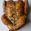 A beautifully roasted chicken sits center of a white marble cutting board. The skin looks crisp and delicious. The herb rub is generously sprinkled across the skin.