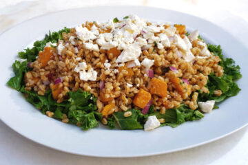 A large oval, family-style platter displays a generous portion of this Easy Butternut Squash Farro Salad with 5-Ingredient Dressing. A bed of fresh, crisp kale frames the salad. Butternut squash, red onion, fresh parsley and feta crumbles invite you to dive in.