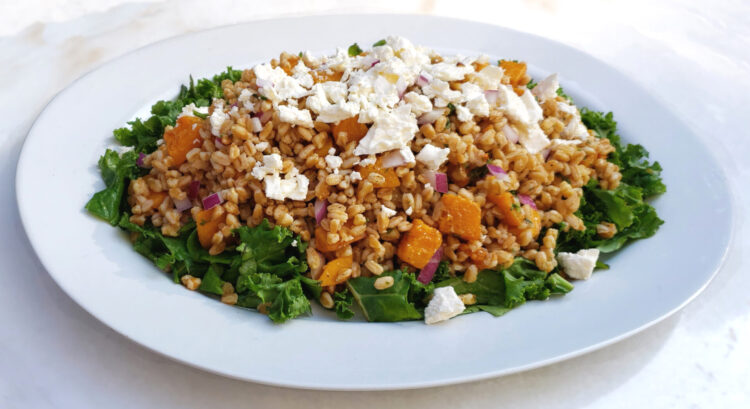 A large oval, family-style platter displays a generous portion of this Easy Butternut Squash Farro Salad with 5-Ingredient Dressing. A bed of fresh, crisp kale frames the salad. Butternut squash, red onion, fresh parsley and feta crumbles invite you to dive in.