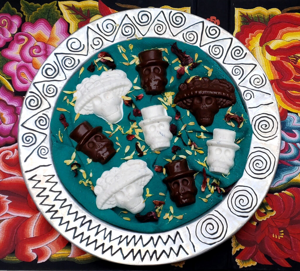 Easy Chocolate Calavera Candies 2 Ways. A rustic, round silver platter with an etched design of swirls and angular images holds a combination of white and dark chocolate skulls. The chocolates are shaped as either elegant ladies wearing elaborate hats or as gentlemen in top hats. The white chocolate candies have toasted coconut and crushed hibiscus flowers mixed within and sprinkled on the backs of the candies, while the dark chocolate candies have roasted pepitas and dried cranberries.