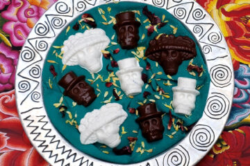 Easy Chocolate Calavera Candies 2 Ways. A rustic, round silver platter with an etched design of swirls and angular images holds a combination of white and dark chocolate skulls. The chocolates are shaped as either elegant ladies wearing elaborate hats or as gentlemen in top hats. The white chocolate candies have toasted coconut and crushed hibiscus flowers mixed within and sprinkled on the backs of the candies, while the dark chocolate candies have roasted pepitas and dried cranberries.