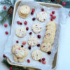 A tray of fresh baked Lemon Cranberry Slice Cookies rests on top of a cool, white marble counter top that looks like snow. Fresh cranberries and lemon zest are sprinkled over the cookies Fresh evergreen sprigs and pine cones frame the rectangular pan of cookies.