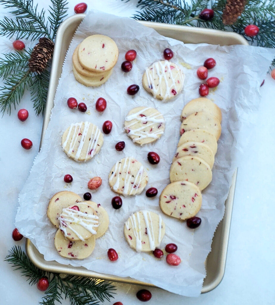 A tray of fresh baked Lemon Cranberry Slice Cookies rests on top of a cool, white marble counter top that looks like snow. Fresh cranberries and lemon zest are sprinkled over the cookies Fresh evergreen sprigs and pine cones frame the rectangular pan of cookies.