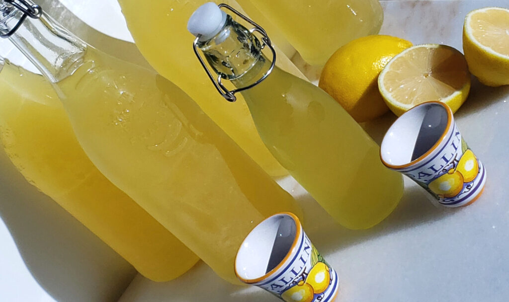 Bottles of homemade Italian Limoncello are lined up on a cool marble countertop. Sunshine and fresh lemons lend a warm glow to the arrangement.