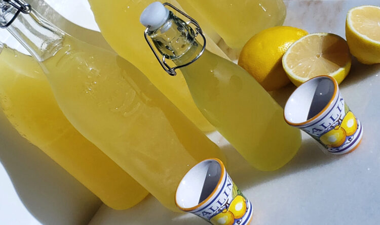 Bottles of homemade Italian Limoncello are lined up on a cool marble countertop. Sunshine and fresh lemons lend a warm glow to the arrangement.