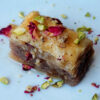 A single piece of Simple 4-Step Cardamom Rose Baklava sits in the center of a plate. Dried rose petals and pistachios are sprinkled on top of this layered dessert.