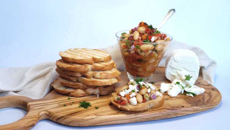 A rustic wooden charcuterie board features the following from left to right: a stack of toasted Italian bread, a small glass bowl of roasted red pepper and cannellini bruschetta and a creamy slice of fresh goat cheese. In the forefront of the picture rests a piece of crusty bread piled high with bruschetta and topped with goat cheese crumbles.