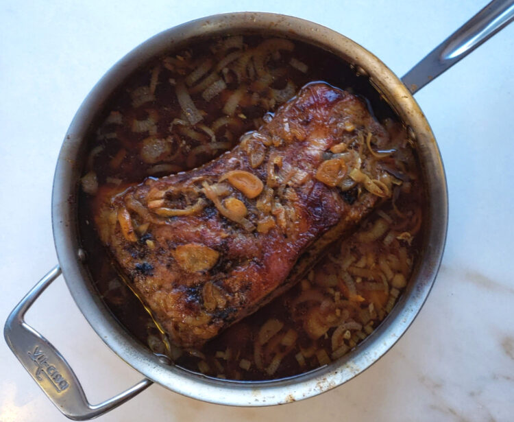 A beautifully braised brisket with Shallots and a rich, beef au jus, sits prominetly in the center of an All Clad Roasting pan. The meat is fork tender.