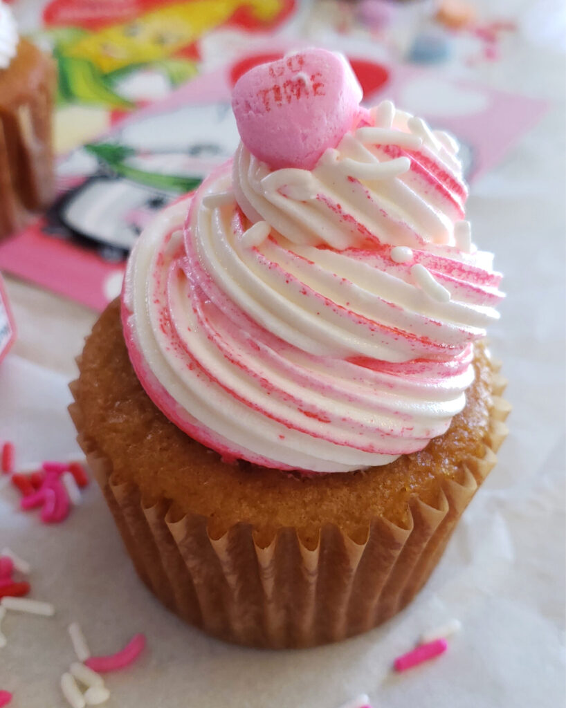 A super moist Easy Brown Sugar Love Cupcake sits front and center. Topped with a swirl of light buttercream, sprinkles and a candy Valentine's Day heart.