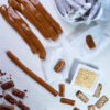 A bright work surface is covered with 6-Ingredient Easy Homemade Caramels in various stages of completion. Some are rolled and cut. Others pieces have been rolled in toasted sesame seeds and dried rose petals for an elegant finish. Others are shown wrapped in candy wrappers.