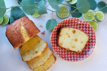 Fresh greens and sliced limes frame a loaf of ginger lime pound cake.Three slices lay stacked on top of each other directly in fromt of the half sliced loaf. A single slice sits centered on a honeycomb textured plate. Pieces of candied ginger and lime zest can be seen throughout the bake.