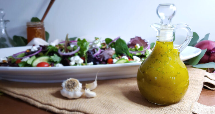 A decanter of Bright Herb and White Balsamic Vinaigrette sits in the forefront of the image. Behind it is a larve oval platter heaped high with a fresh salad consisting of mixed baby greens, crisp cucumbers, sweet tomatoes, red onions. Goat cheese crumbles, toasted pepitas and dried cherries are sprinkled across the top. the fresh herbs and garlic are perfectly suspended in this golden dressing.