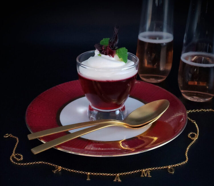 An elegant glass of Easy 2-Step Hibiscus Flower and Prosecco Gelee sits in the center of a fine china serving plate. Two spoons nest together across the plate, suggesting this dessert is meant to be shared. Two stemless champaigne flutes are bubbling in the background. A gold necklace spells out the words Ti Amo (I love you) in Italian across the front of the image.
