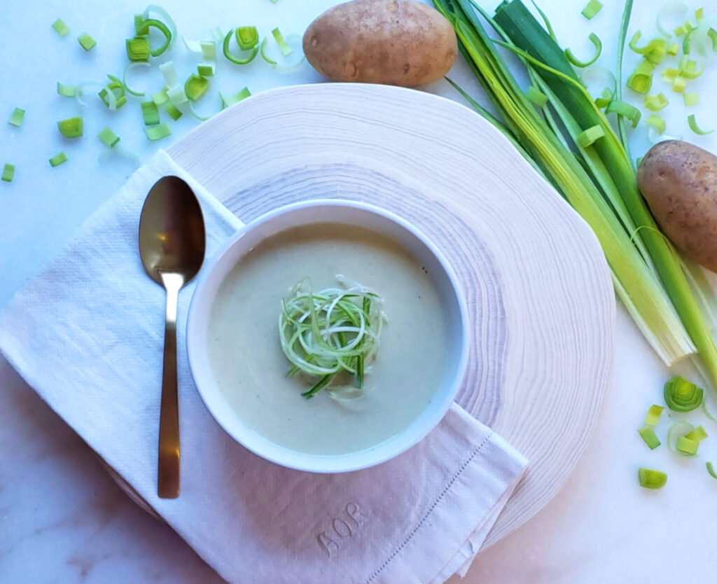 Fresh leeks and new potatoes surround a hot bowl of this Easy 30-Minute Creamy Potato Leek Soup. A simple bowl rests upon a rustic, round board from a cross section of a tree. The growth circles from the tree radiate outward, framing the bowl perfectly. Upon an antique napkin rests a simple spoon. The picture has a very natural, organic and wholesome appearance.