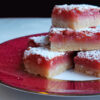 A close up shot of a stack of Tempting 2 Bite Blood Orange Bars. The shortbread crust shows flecks of coconut. Blood Orange zest is sprinkled throughout the filling and the top of the bar is dusted with powdered sugar.