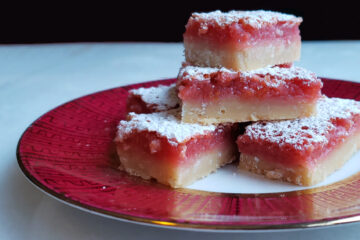 A close up shot of a stack of Tempting 2 Bite Blood Orange Bars. The shortbread crust shows flecks of coconut. Blood Orange zest is sprinkled throughout the filling and the top of the bar is dusted with powdered sugar.