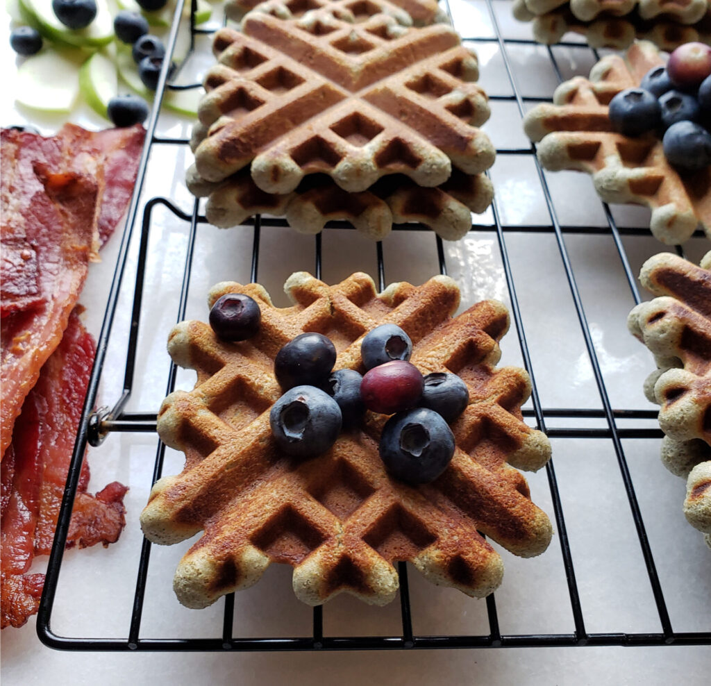 Rows of Easy 15-Minute Blue Corn Buttermilk Waffles are lined up on a marble board. Crispy thick cut bacon is shown on the left side, along with fresh blueberries and thinly sliced granny smith apples. A waffle with fresh blueberries sits front and center of the image - tempting you. A stack of waffles is generously  topped with this Easy 20-Minute Thyme Infused Blueberry Sauce. 