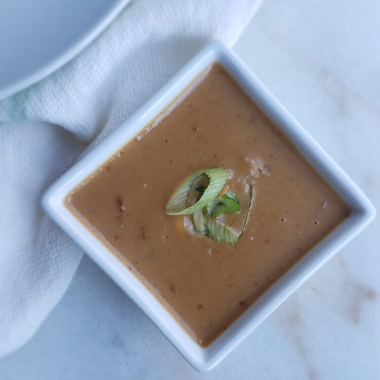 A cup of deliciously creamy Easy 15-minute Thai Peanut Coconut Sauce is topped with fresh, sliced scallions.